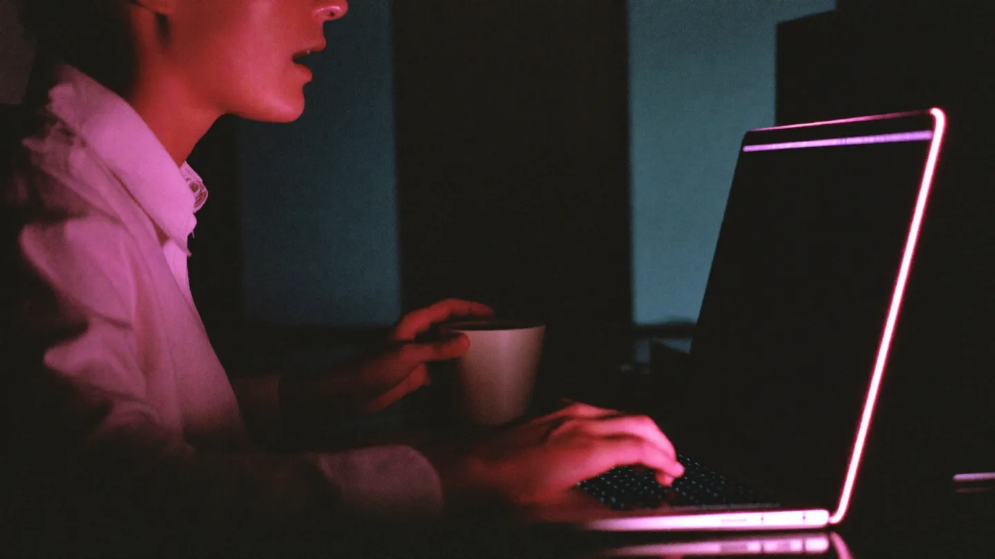 Someone at a desk at night, speaking toward an open MacBook whose screen is dark except for a small glowing pink strip under the camera.