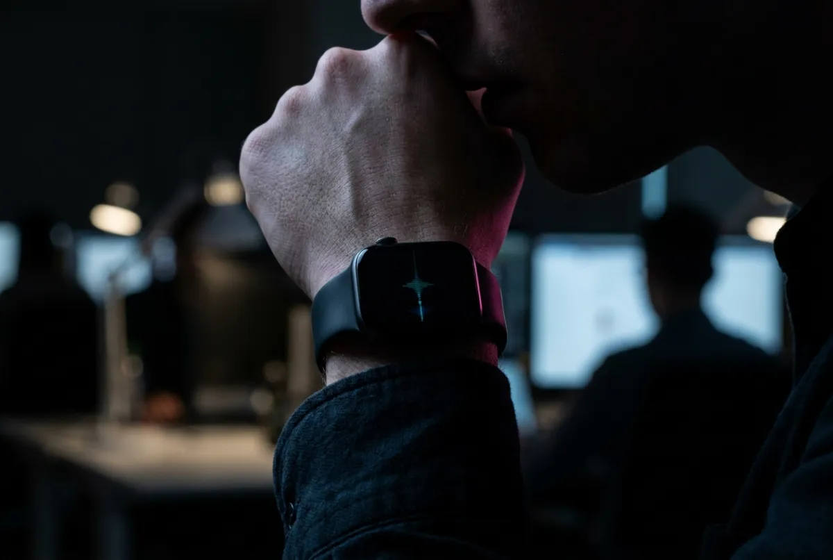 Close-up of someone in a dim open-plan office holding their wrist to their mouth, speaking quietly into an Apple Watch whose screen shows a live audio waveform.